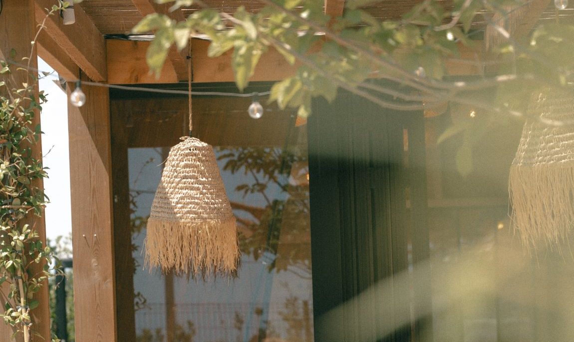 La terrasse d'ORA vue de l'extérieur à travers le feuillage : pergola en bois et suspension en raphia